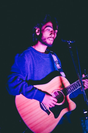 A person is playing an acoustic guitar under stage lighting, wearing a casual long-sleeved shirt. The lighting casts a purple hue on the scene, highlighting the musician's face and instrument.