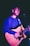 A person is playing an acoustic guitar under stage lighting, wearing a casual long-sleeved shirt. The lighting casts a purple hue on the scene, highlighting the musician's face and instrument.