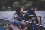 A person with long hair plays an acoustic guitar and sings into a microphone. Another individual sits nearby, listening attentively. They are outdoors, possibly near a body of water, with greenery in the background. The scene appears to be lit by artificial light, casting soft shadows.