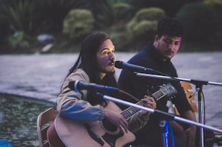 A person with long hair plays an acoustic guitar and sings into a microphone. Another individual sits nearby, listening attentively. They are outdoors, possibly near a body of water, with greenery in the background. The scene appears to be lit by artificial light, casting soft shadows.