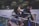 A person with long hair plays an acoustic guitar and sings into a microphone. Another individual sits nearby, listening attentively. They are outdoors, possibly near a body of water, with greenery in the background. The scene appears to be lit by artificial light, casting soft shadows.