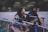 A person with long hair plays an acoustic guitar and sings into a microphone. Another individual sits nearby, listening attentively. They are outdoors, possibly near a body of water, with greenery in the background. The scene appears to be lit by artificial light, casting soft shadows.