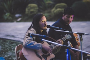 A person with long hair plays an acoustic guitar and sings into a microphone. Another individual sits nearby, listening attentively. They are outdoors, possibly near a body of water, with greenery in the background. The scene appears to be lit by artificial light, casting soft shadows.