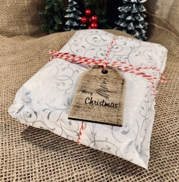 A neatly wrapped gift, adorned with decorative white and silver swirl-patterned paper, is tied with red and white twine. A brown wooden gift tag that reads 'Merry Christmas' with a simple tree design is attached to the twine. In the background, there are small decorated Christmas trees with red baubles and frosted branches, set against a textured burlap surface.