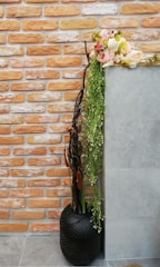 A decorative arrangement featuring a modern black vase with perforations, containing tall branches, cattails, and cascading greenery. The top is adorned with a lush bouquet of pink and white flowers. The background consists of a wall with orange and white bricks next to a smooth, gray surface, adding contrast to the vibrant floral display.