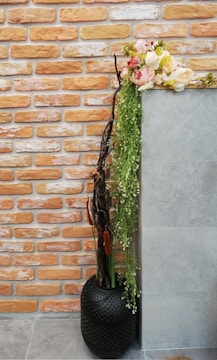 A decorative arrangement featuring a modern black vase with perforations, containing tall branches, cattails, and cascading greenery. The top is adorned with a lush bouquet of pink and white flowers. The background consists of a wall with orange and white bricks next to a smooth, gray surface, adding contrast to the vibrant floral display.