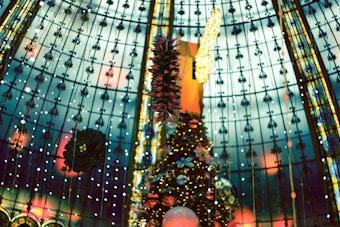 A decorated indoor setting featuring a large Christmas tree adorned with ornaments and lights. The ceiling is made of glass panels with intricate patterns and hanging decorations which include wreaths and spiral ornaments. Soft warm lights accent the overall ambience.