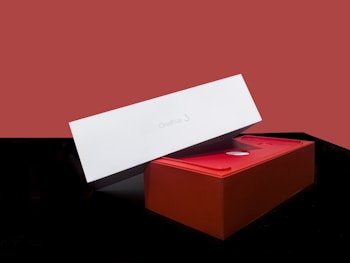 A red and white rectangular box partially open, revealing the interior. The box has the text 'OnePlus 3' printed on the top of the white lid. The background is a solid muted red, and the surface on which the box rests is black.