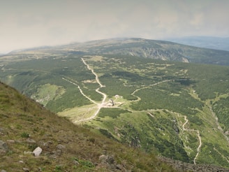 A lush green landscape with wide open spaces and rolling hills, intersected by a winding dirt path that stretches into the distance. A building complex is nestled in a clearing surrounded by greenery, and in the distance, a range of gentle mountains fades into the horizon under a cloudy sky.