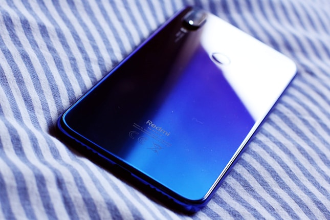 A smartphone with a shiny gradient back cover featuring a dark blue to light blue transition. It is placed on a soft, striped fabric surface.