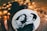 A black and white embroidered portrait of a couple in a circular hoop placed on a textured surface. Surrounding the embroidery are small, warm yellow fairy lights and dark brown pine cones. A glossy, brown leaf lies nearby, enhancing the rustic and cozy atmosphere.