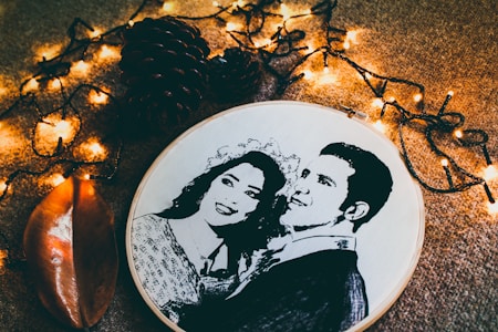 A black and white embroidered portrait of a couple in a circular hoop placed on a textured surface. Surrounding the embroidery are small, warm yellow fairy lights and dark brown pine cones. A glossy, brown leaf lies nearby, enhancing the rustic and cozy atmosphere.