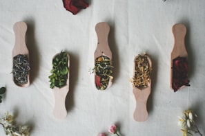 Five wooden scoops are arranged in a row on a light fabric background, each filled with different dried herbs and flowers. The selection includes lavender, green herbs, rose petals, and other floral elements. Surrounding the scoops are scattered petals and small flowers, contributing to a rustic and natural aesthetic.