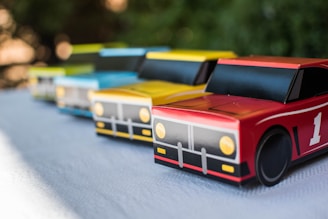 Four toy race cars in vibrant colors are lined up on a textured surface. The cars feature simplistic designs with distinct details, including large wheels and printed headlights. They are colored in shades of red, yellow, blue, and green, arranged diagonally from the front to the back.