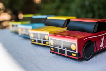 Four toy race cars in vibrant colors are lined up on a textured surface. The cars feature simplistic designs with distinct details, including large wheels and printed headlights. They are colored in shades of red, yellow, blue, and green, arranged diagonally from the front to the back.