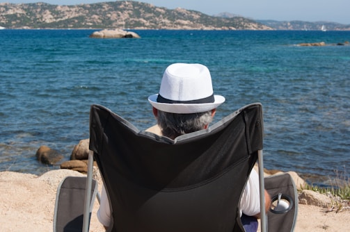 A person wearing a white hat is sitting in a black folding chair facing the sea. The scene includes clear blue water, a rocky coastline, and distant hills under a bright sky.