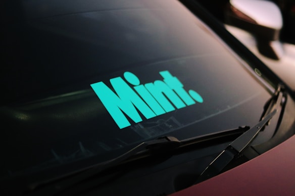 A car windshield with a bold mint-colored sticker displaying the word 'Mint.' The windshield wipers are visible, and there is a dark ambiance surrounding the car.