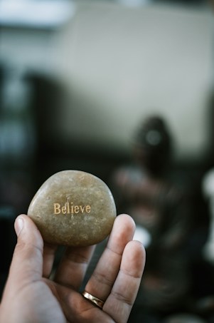 A hand is holding a smooth stone with the word 'Believe' engraved on it. The background is slightly blurred, hinting at an indoor setting with dim lighting and a possibly decorative item in the background.