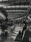 A spacious, modern airport terminal features a high, arched ceiling with geometric metal frames. In the foreground, a person leans against a structure while using their phone, with a suitcase positioned nearby. People carrying luggage move through the terminal, creating a sense of motion blur. The terminal is illuminated by natural light streaming through large windows, casting patterns of light and shadow on the floor.