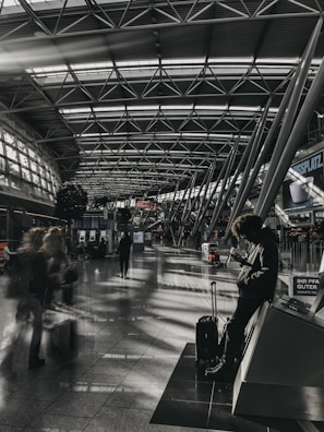 A spacious, modern airport terminal features a high, arched ceiling with geometric metal frames. In the foreground, a person leans against a structure while using their phone, with a suitcase positioned nearby. People carrying luggage move through the terminal, creating a sense of motion blur. The terminal is illuminated by natural light streaming through large windows, casting patterns of light and shadow on the floor.