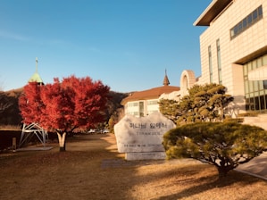 A large stone with Korean writing and 'CORAM DEO' inscribed on it lies on a manicured lawn. To the left, a vibrant red tree stands near a structure with a cross on top. In the background, a building with modern architectural features and tall windows is visible, alongside carefully trimmed greenery.