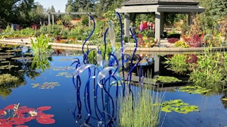 An outdoor garden scene with a serene pond, featuring a variety of colorful plants and flowers around its edges. In the center of the pond, a striking glass sculpture with wavy blue and translucent strands adds an artistic touch. A gazebo structure with people inside is visible in the background, surrounded by more vibrant foliage and plant life.