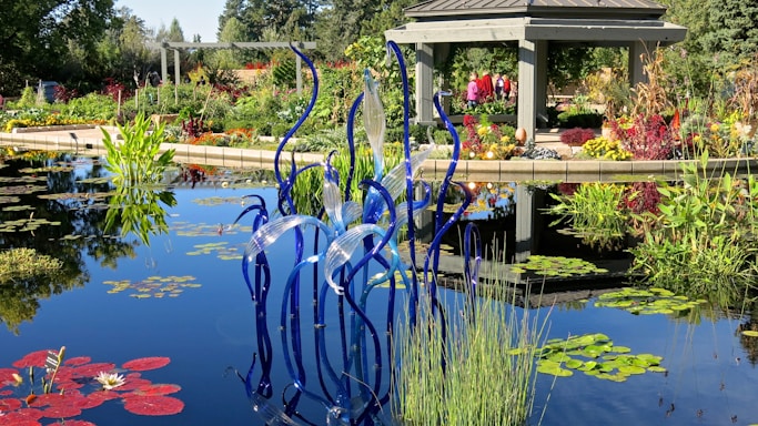 An outdoor garden scene with a serene pond, featuring a variety of colorful plants and flowers around its edges. In the center of the pond, a striking glass sculpture with wavy blue and translucent strands adds an artistic touch. A gazebo structure with people inside is visible in the background, surrounded by more vibrant foliage and plant life.