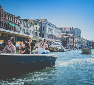 A group of people are enjoying a boat ride along a canal surrounded by historic buildings. The architecture includes a blend of stone facades and vibrant paint, typical of a European city. The canal is bustling with various boats and the sky is clear with a bright blue hue.