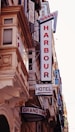 A street view featuring a building façade with prominent retro-style signs. The signs include 'GRAND HARBOUR' in large vertical letters and 'HOTEL' with an arrow pointing toward the entrance. The architecture includes ornate details and numerous windows with shutters.