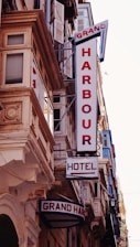 A street view featuring a building façade with prominent retro-style signs. The signs include 'GRAND HARBOUR' in large vertical letters and 'HOTEL' with an arrow pointing toward the entrance. The architecture includes ornate details and numerous windows with shutters.