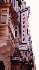 A street view featuring a building façade with prominent retro-style signs. The signs include 'GRAND HARBOUR' in large vertical letters and 'HOTEL' with an arrow pointing toward the entrance. The architecture includes ornate details and numerous windows with shutters.