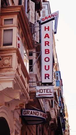 A street view featuring a building façade with prominent retro-style signs. The signs include 'GRAND HARBOUR' in large vertical letters and 'HOTEL' with an arrow pointing toward the entrance. The architecture includes ornate details and numerous windows with shutters.