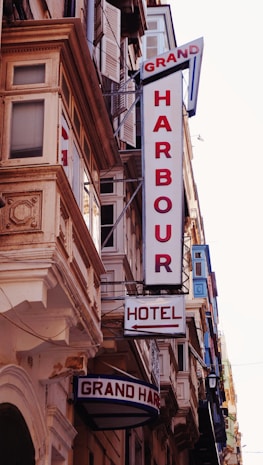 A street view featuring a building fa&ccedil;ade with prominent retro-style signs. The signs include 'GRAND HARBOUR' in large vertical letters and 'HOTEL' with an arrow pointing toward the entrance. The architecture includes ornate details and numerous windows with shutters.
