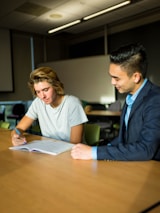 Two individuals are seated at a wooden table in a classroom or office setting. One person, wearing a gray t-shirt, is writing in a notebook, while the other, dressed in a suit, observes and offers guidance. The room appears to have minimal lighting, with a soft natural light coming through the windows.