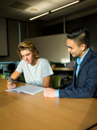 Photograph of a friendly mentor advising a student over documents in a modern study setting.