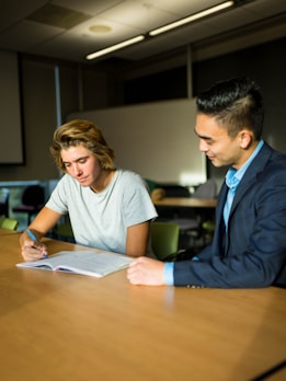 Two individuals are seated at a wooden table in a classroom or office setting. One person, wearing a gray t-shirt, is writing in a notebook, while the other, dressed in a suit, observes and offers guidance. The room appears to have minimal lighting, with a soft natural light coming through the windows.