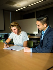 Two individuals are seated at a wooden table in a classroom or office setting. One person, wearing a gray t-shirt, is writing in a notebook, while the other, dressed in a suit, observes and offers guidance. The room appears to have minimal lighting, with a soft natural light coming through the windows.