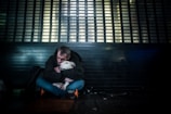 A person sits on the ground in a dimly lit environment, embracing a white dog. They both appear close to a metallic or barred structure, hinting at a cityscape setting. The atmosphere feels somber and intimate.