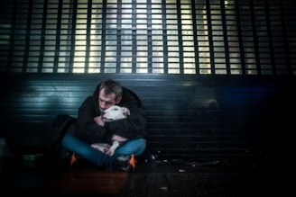 A person sits on the ground in a dimly lit environment, embracing a white dog. They both appear close to a metallic or barred structure, hinting at a cityscape setting. The atmosphere feels somber and intimate.