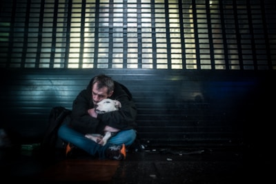 A person sits on the ground in a dimly lit environment, embracing a white dog. They both appear close to a metallic or barred structure, hinting at a cityscape setting. The atmosphere feels somber and intimate.