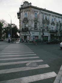 A street scene with an old, multi-story building at the corner. The building has a worn facade and multiple windows with decorative elements. In front of the building, there is a pedestrian crosswalk leading towards a shop with a sign labeled 'Mega Image.' Trees and several pedestrians are visible along the sidewalk, and some vehicles are parked or moving nearby.