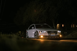 A white car with its headlights on, parked or driving on a dimly lit road at night. The license plate is visible, and surrounding trees and buildings are in shadow, adding to the nighttime ambiance.