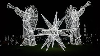 Two large angel figures made of white lights holding trumpets stand on either side of a bright star-shaped light installation on a dark background. The scene is illuminated by these lights, casting a glowing effect and creating a festive atmosphere.