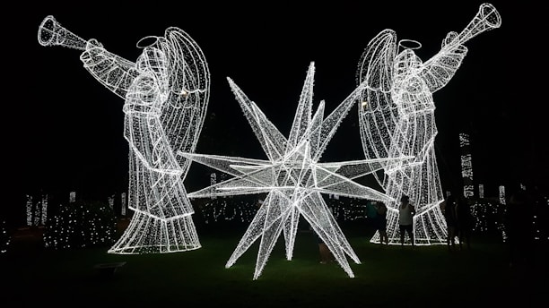 Two large angel figures made of white lights holding trumpets stand on either side of a bright star-shaped light installation on a dark background. The scene is illuminated by these lights, casting a glowing effect and creating a festive atmosphere.