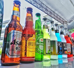 A variety of bottled beverages are displayed in a neat row on a reflective glass surface. The bottles, of various shapes and sizes, are filled with different colored liquids, featuring diverse labels from various brands. The setting appears to be outdoors with a metal beam structure overhead, suggesting a possible event or market environment.