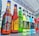 A variety of bottled beverages are displayed in a neat row on a reflective glass surface. The bottles, of various shapes and sizes, are filled with different colored liquids, featuring diverse labels from various brands. The setting appears to be outdoors with a metal beam structure overhead, suggesting a possible event or market environment.
