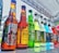 A variety of bottled beverages are displayed in a neat row on a reflective glass surface. The bottles, of various shapes and sizes, are filled with different colored liquids, featuring diverse labels from various brands. The setting appears to be outdoors with a metal beam structure overhead, suggesting a possible event or market environment.