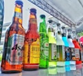 A variety of bottled beverages are displayed in a neat row on a reflective glass surface. The bottles, of various shapes and sizes, are filled with different colored liquids, featuring diverse labels from various brands. The setting appears to be outdoors with a metal beam structure overhead, suggesting a possible event or market environment.