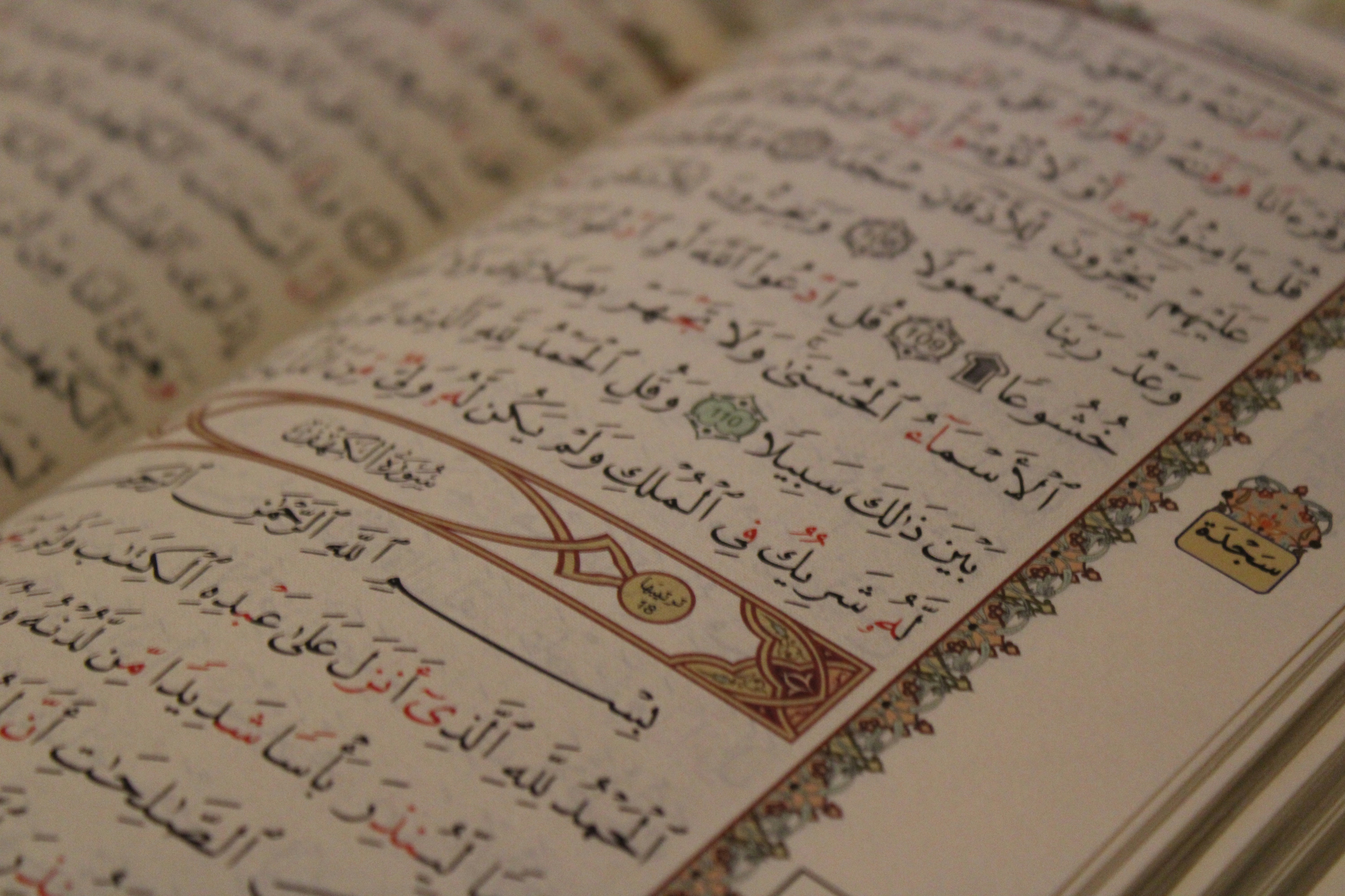 An open Arabic book displaying intricate calligraphy with decorative elements on the margins. The text is organized into lines with a mix of black and red script, and features ornamental designs and symbols.