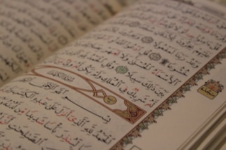 An open Arabic book displaying intricate calligraphy with decorative elements on the margins. The text is organized into lines with a mix of black and red script, and features ornamental designs and symbols.